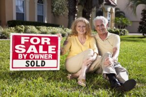 Senior couple sitting on the grass outside their home with For Sale Sold sign