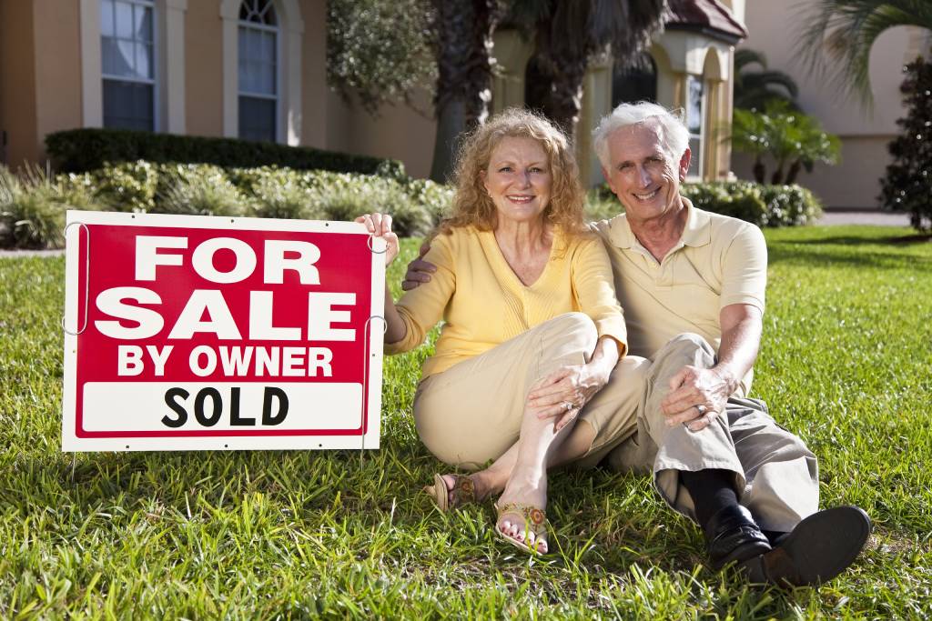 Senior couple sitting on the grass outside their home with For Sale Sold sign